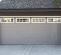 Gray residential garage door with rectangular panel design and a row of small square windows at the top, set in a modern house exterior.