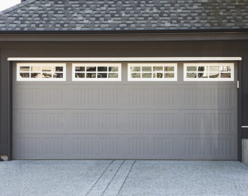 Gray residential garage door with rectangular panel design and a row of small square windows at the top, set in a modern house exterior.