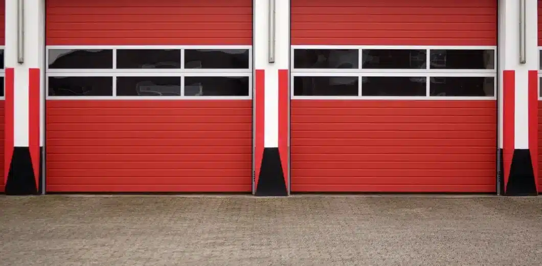 Two closed red garage doors with horizontal black and white stripes, set in a white and red building, viewed from a concrete driveway.