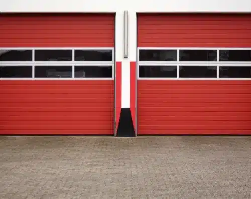 Two closed red garage doors with horizontal black and white stripes, set in a white and red building, viewed from a concrete driveway.