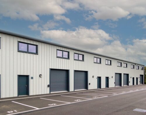 A row of modern industrial units with large roller doors and parking spaces marked in front, under a partly cloudy sky.