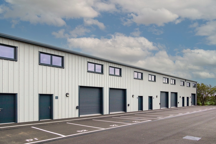 A row of modern industrial units with large roller doors and parking spaces marked in front, under a partly cloudy sky.