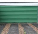 Closed green garage door with horizontal panels, set between two brick walls, with a gravel and concrete driveway in front.