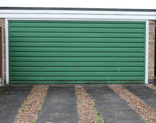 Closed green garage door with horizontal panels, set between two brick walls, with a gravel and concrete driveway in front.