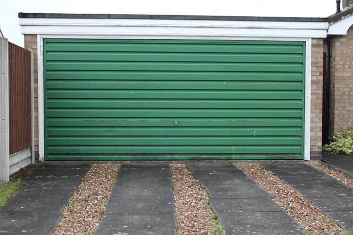 Closed green garage door with horizontal panels, set between two brick walls, with a gravel and concrete driveway in front.