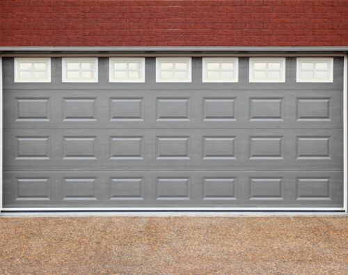 A closed gray garage door with small rectangular windows at the top, set in a red brick wall with two outdoor wall lights on either side.