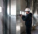 A worker in a white hard hat uses a power tool to work on a metal ventilation duct inside an industrial building.