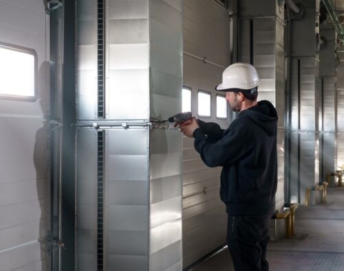 A worker in a white hard hat uses a power tool to work on a metal ventilation duct inside an industrial building.
