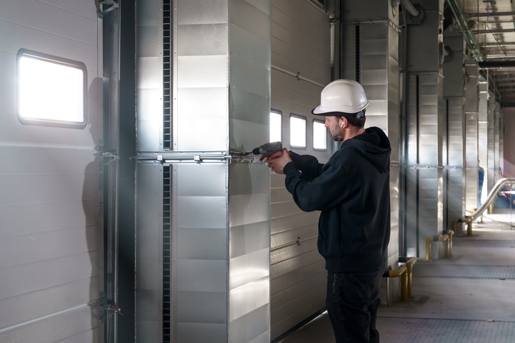 A worker in a white hard hat uses a power tool to work on a metal ventilation duct inside an industrial building.