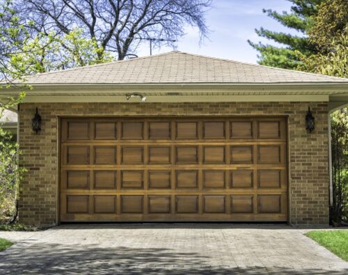A brick house with a closed wooden garage door, two outdoor wall lanterns, and a paved driveway surrounded by trees and shrubs.