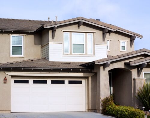Two-story suburban house with beige stucco exterior, white trim, a double garage door, and landscaped front yard.
