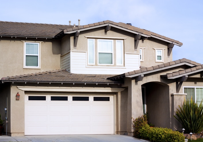 Two-story suburban house with beige stucco exterior, white trim, a double garage door, and landscaped front yard.