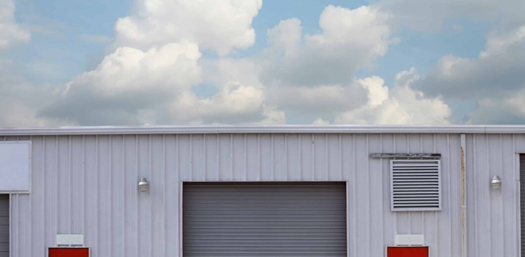 A white industrial building with a large gray garage door centered between two bright red doors, under a partly cloudy sky.