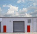 A white industrial building with a large gray garage door centered between two bright red doors, under a partly cloudy sky.
