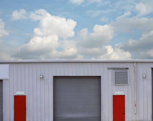 A white industrial building with a large gray garage door centered between two bright red doors, under a partly cloudy sky.