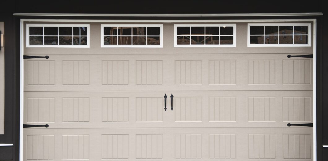 Closed beige garage door with rectangular panel design and four windows at the top, featuring black decorative handles and hardware.