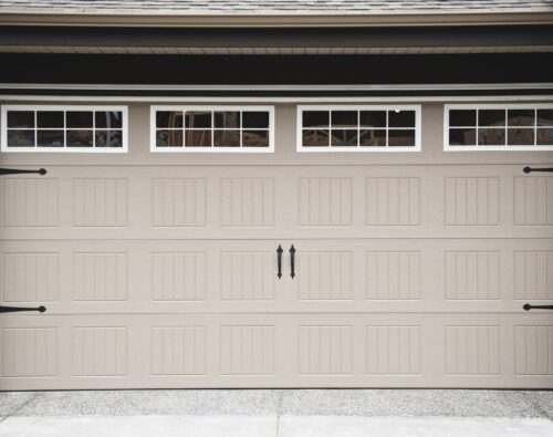 Closed beige garage door with rectangular panel design and four windows at the top, featuring black decorative handles and hardware.