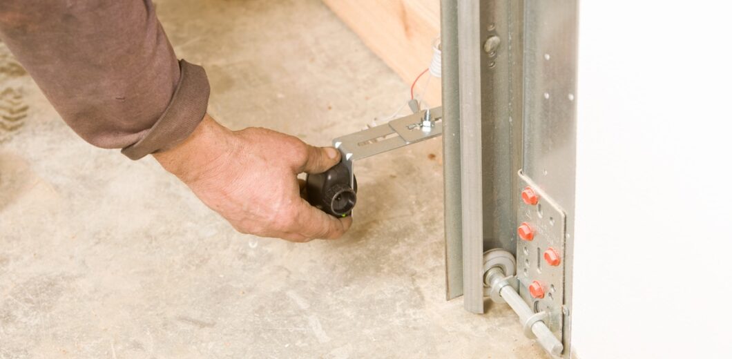 A person’s hand adjusts a metal component on a garage door track near the concrete floor.