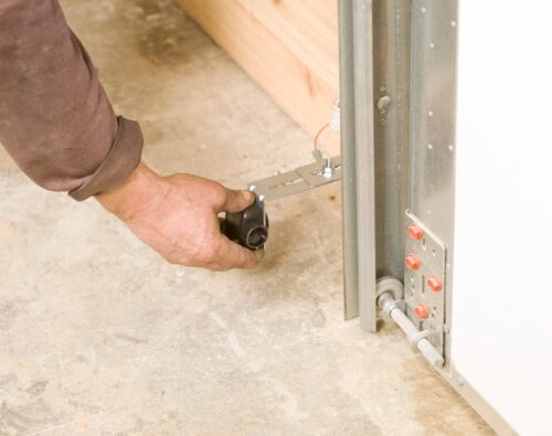 A person’s hand adjusts a metal component on a garage door track near the concrete floor.