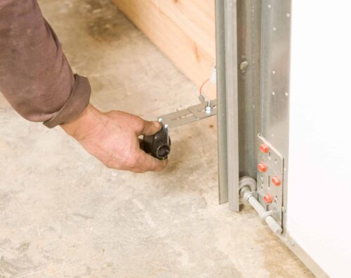 A person adjusts the sensor near the bottom of a garage door track, with visible wiring and mounting hardware on a concrete floor.