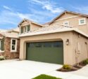 Two-story beige house with a green garage door, stone accents, and a manicured front yard under a blue sky.