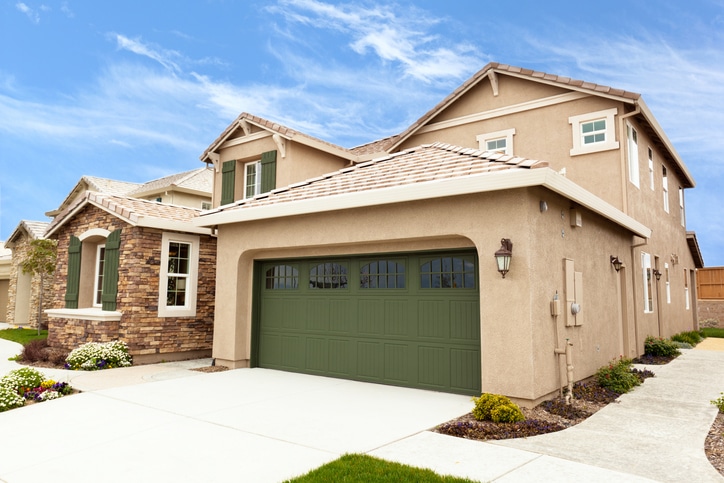 Two-story beige house with a green garage door, stone accents, and a manicured front yard under a blue sky.