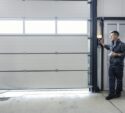 A man in work clothes stands by a control panel next to a large closed garage door in an industrial setting.