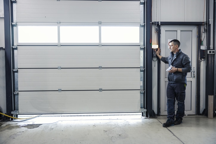A man in work clothes stands by a control panel next to a large closed garage door in an industrial setting.