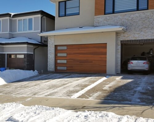A modern two-story house with a wooden garage door and a car parked inside the garage. The driveway and sidewalk are partially cleared of snow.