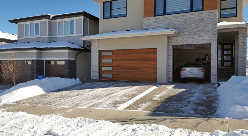 A modern two-story house with a wooden garage door and a car parked inside the garage. The driveway and sidewalk are partially cleared of snow.