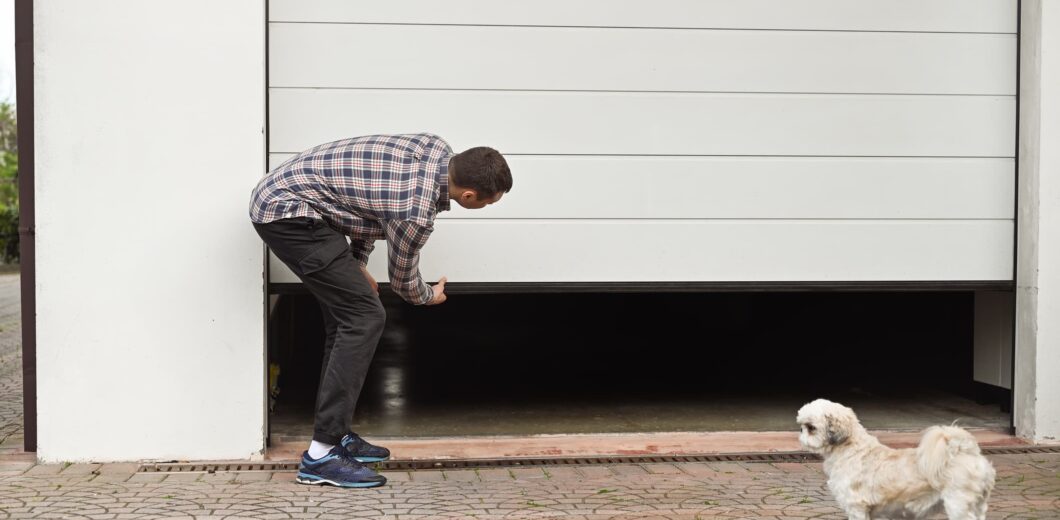 A man in a plaid shirt manually lifts a white garage door while a small white dog stands nearby on a brick driveway.