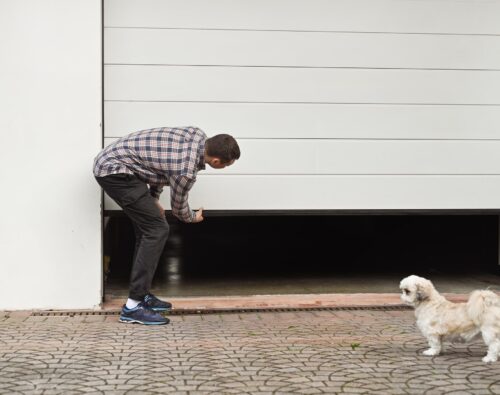A man in a plaid shirt manually lifts a white garage door while a small white dog stands nearby on a brick driveway.