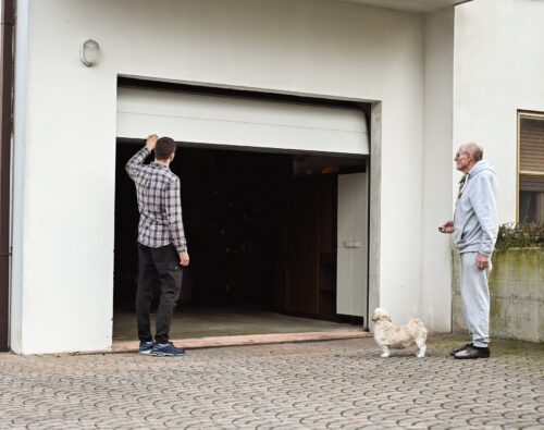 Two men and a small dog stand outside an open garage door attached to a white building. One man is facing the garage, and the other is standing nearby with the dog.