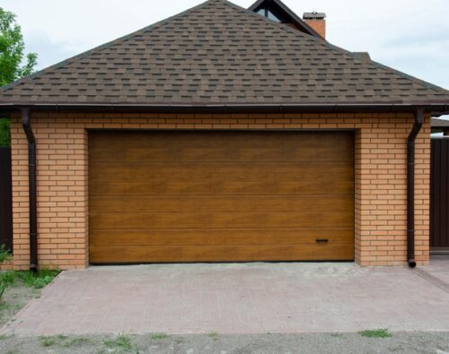 A closed wooden garage door set in a brick-walled garage with a paved driveway in front.