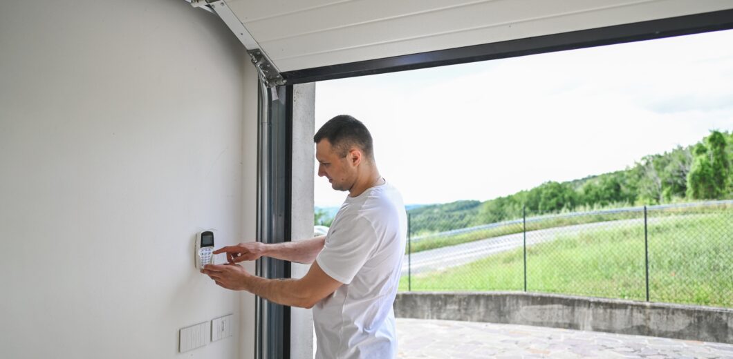 A man in a white t-shirt enters a code on a wall-mounted keypad inside a garage with the door open, overlooking a paved driveway and greenery outside.