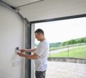 A man in a white t-shirt enters a code on a wall-mounted keypad inside a garage with the door open, overlooking a paved driveway and greenery outside.