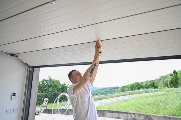 A person in a white shirt adjusts or repairs the inside of a partially open garage door during the daytime.
