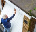 An older man with gray hair is cleaning or repairing a white garage door attached to a brick house, with moss visible on the roof edge.