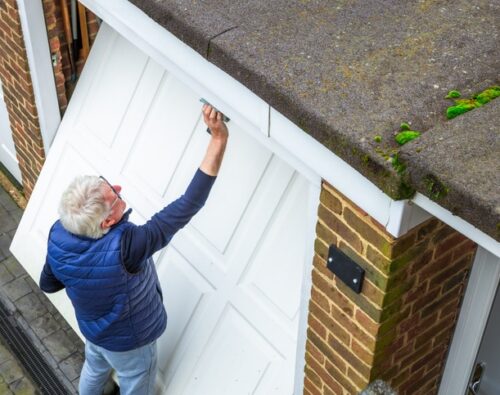 An older man with gray hair is cleaning or repairing a white garage door attached to a brick house, with moss visible on the roof edge.