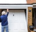 An older person in a blue vest wipes the top of a white garage door that is partially open, with a bucket nearby on the ground.