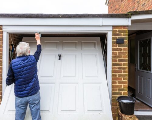 An older person in a blue vest wipes the top of a white garage door that is partially open, with a bucket nearby on the ground.