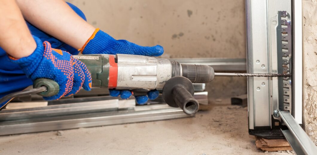 A person wearing blue gloves uses a power drill to bore a hole into a metal frame at a construction site.