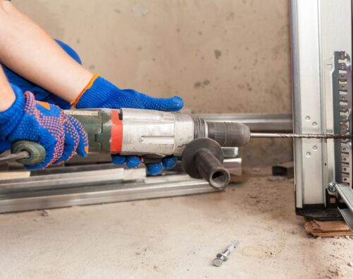 A person wearing blue gloves uses a power drill to bore a hole into a metal frame at a construction site.