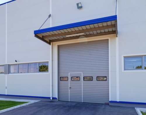 Gray industrial garage door with small windows in a white commercial building, surrounded by pavement and green grass.
