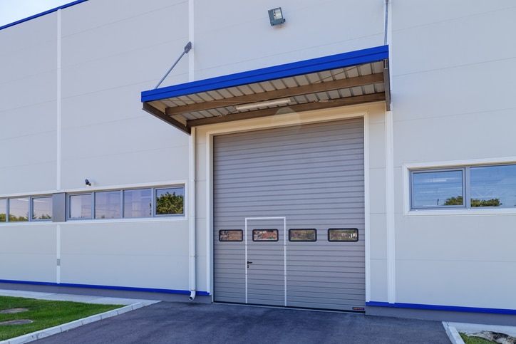 Gray industrial garage door with small windows in a white commercial building, surrounded by pavement and green grass.
