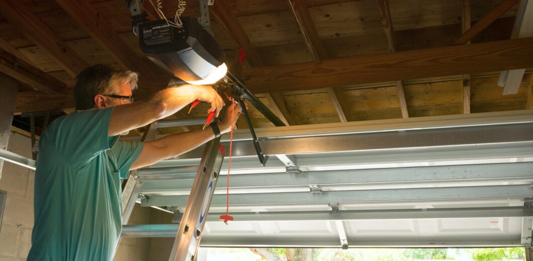 A man standing on a ladder repairs a garage door opener in a residential garage with the door partially open.