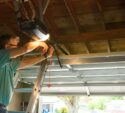 A man standing on a ladder repairs a garage door opener in a residential garage with the door partially open.