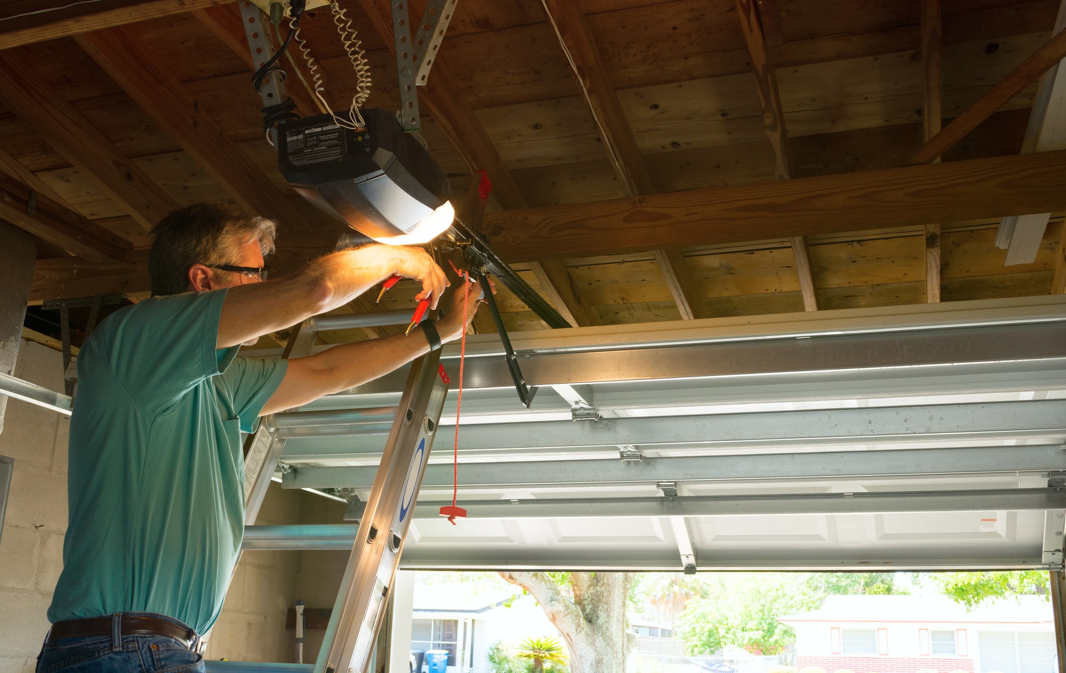 A man standing on a ladder repairs an automatic garage door opener inside a garage with the door partially open.