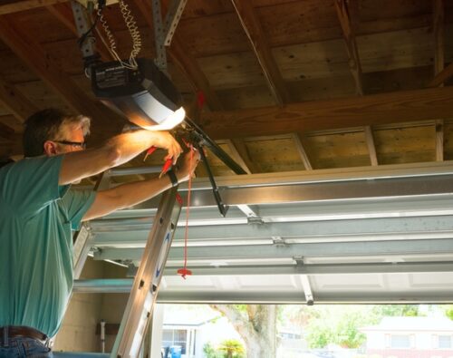 A person stands on a ladder adjusting a garage door opener mechanism inside a partially open garage.