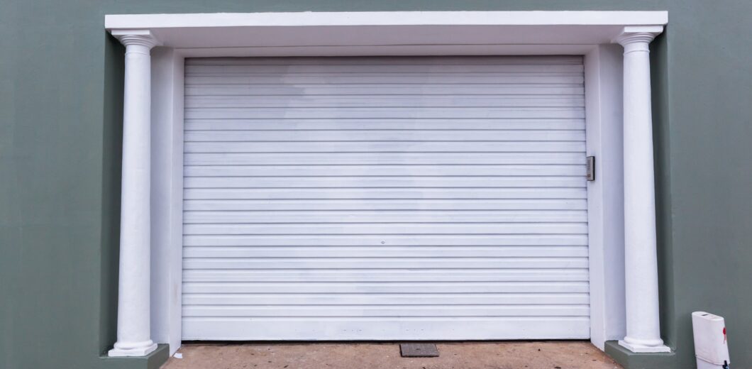 A closed white garage door with vertical ridges, framed by two white columns against a green wall. Concrete driveway in front.